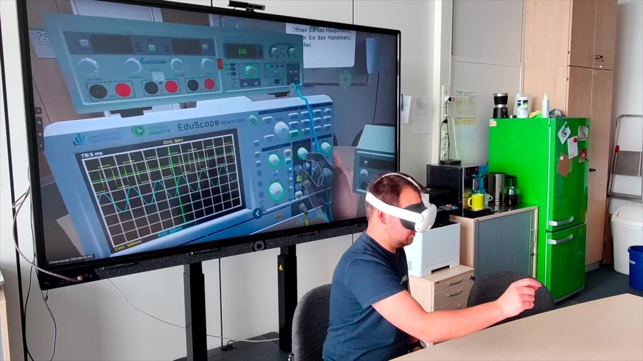 Ein Mann sitzt mit einer VR-Brille an einem Tisch und greift vor ihm in die Luft. Hinter ihm steht ein großer Bildschirm, auf dem eine VR-Abbildung gezeigt wird.