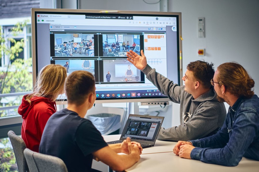 Eine Gruppe von Studierenden sitzen am Tisch und diskutieren vor einem größeren Bildschirm, auf dem verschiedene Aufzeichnungen von Lehr-Szenarien zu sehen sind..