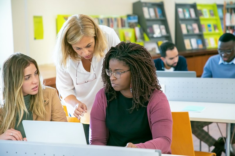 Zwei Studierende sitzen vor dem PC. Hinter ihnen beugt sich die Dozentin über die beiden und erklärt etwas.