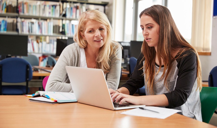 Betreuung bei wissenschaftlichen Arbeiten Zwei Frauen sitzen am Tisch in einer Bibliothek, die Jüngere tippt etwas in ihren Laptop, der vor ihr steht. Die ältere Frau neben ihr schaut ihr zugewandt ebenfalls auf den Laptop-Bildschirm.
