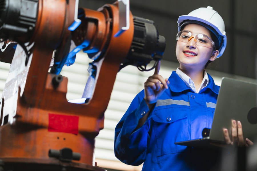 Junge Frau in Arbeitskleidung mit Helm, Brille und Overall, hält Laptop in der Hand und inspiziert eine Maschine.