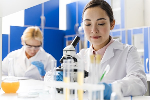 Zwei Wissenschaftlerinnen im Labor. Die Frau im Vordergrund trägt einen weißen Kittel, vor ihr steht ein Mikroskop.