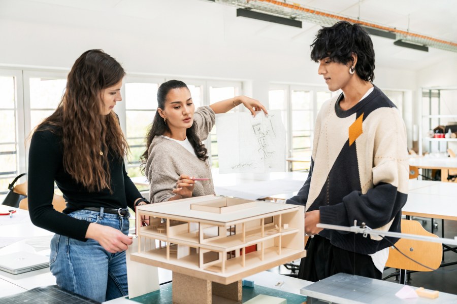 Projektlernen Drei Studierende stehen in einem Seminarraum um ein Modellhaus herum und diskutieren miteinander.