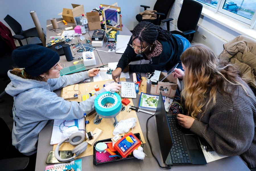 Projektlernen in Gruppen Drei Studierende sind über einen Tisch gebeugt, auf dem viele (Klein-)Teile liegen, u. a. Laptop, Papierblock, Kabel und Werkzeug.