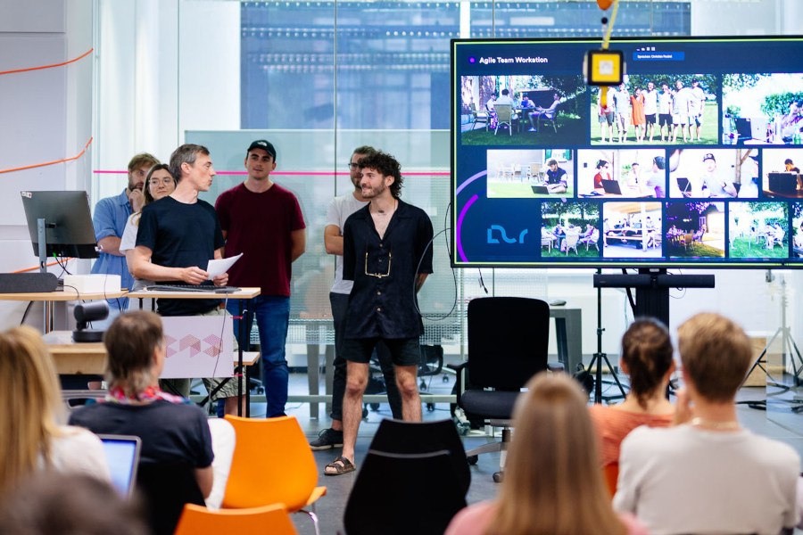 Projektpräsentation Eine Gruppe Studierender hält vor ihren Kommilitoninnen und Kommilitonen eine Präsentation. Neben ihnen steht ein großer Bildschirm, auf dem viele Kacheln zu sehen sind.