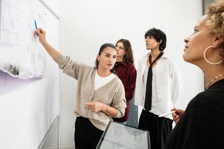 Prüfen im Projektlernen Drei Studierende stehen vor einem Whiteboard, an dem ein Plakat hängt. Eine Studierende zeigt darauf, während sie die Dozierende anschaut, die etwas weiter weg steht und ihr aufmerksam zuhört.