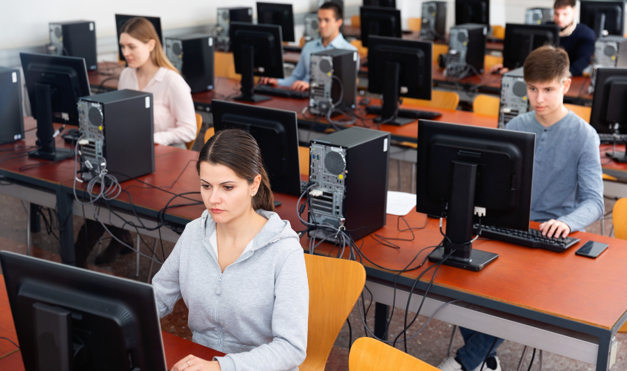 Studierende sitzen konzentriert in einem Raum an Tischen, vor ihren steht jeweils ein Computer.