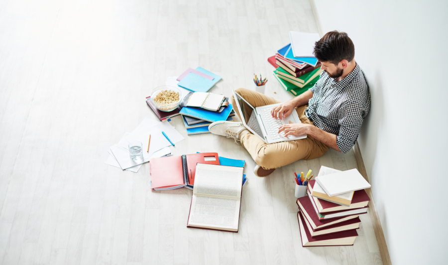 Ein Mann sitzt mit einem Laptop auf dem Schoß auf dem Boden und schreibt. Um ihn herum stapeln sich Bücher und Ordner.