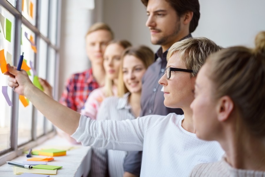 Interaktiver Workshop Sechs Personen schauen gemeinsam auf eine Wand, die mit Post-It-Zettel beklebt ist. Eine Frau mit Brille streckt ihren Arm in Richtung eines Post-It-Zettels, als würde sie ihn gerade hinkleben oder abnehmen.