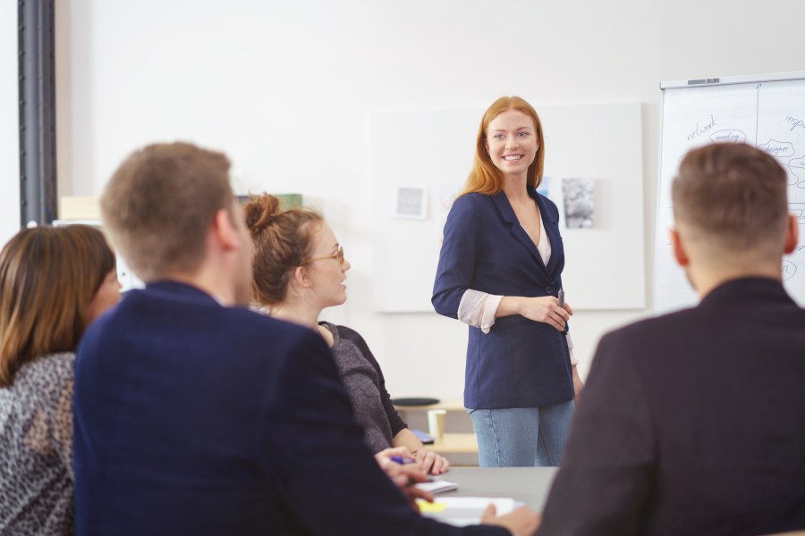 Eine Frau in einem Blazer steht vor einer sitzenden Gruppe von vier Personen. Neben ihr steht ein Flipchart.