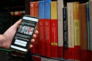 Vor ein Bücherregal wird ein Smartphone gehalten, auf dessen Display eine Quiz-Aufgabe zur Bibliothek zu sehen ist.