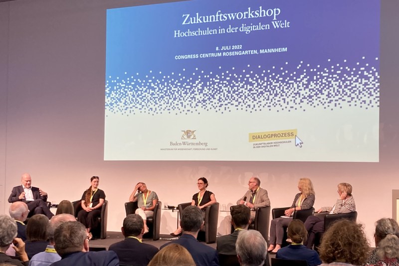 MWK-Dialogprozess Sieben Podiumsgäste sitzen in einem Halbkreis auf der Bühne. Im Hintergrund ist ein Bild an die Wand projiziert, auf dem steht "Zukunftsworkshop Hochschulen in der Digitalen Welt"