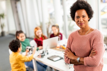 Lehrerin steht vorne rechts mit verschränkten Armen und schaut in die Kamera. Hinter ihr sitzen vier Kinder an einem Tisch, vor ihnen steht ein Laptop. Link zur Themenseite 