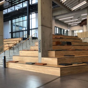 Große Sitz-Tribüne in einer Eingangshalle, unterbrochen von einem Treppenaufgang. Die Sitz-Tribüne aus hellem Holz verläuft über Eck und besteht aus sechs Ebenen. Auf den Ebenen liegen Sitzpolster verstreut. 