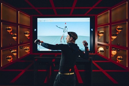 Eindruck aus dem Future Innovation Space (FIS) des IWM Ein Mann ist in einem Raum voller VR-Brillen und hat eine bestimmte Körperposition eingenommen, als würde er gleich etwas werfen. Hinter ihm ist ein Bildschirm zu sehen, der eine abstrakte Person zeigt, die die gleiche Position wie der Mann einnimmt.