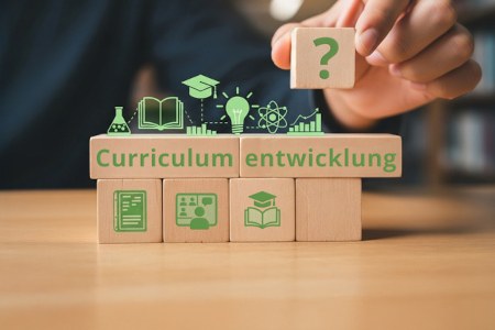 Themenspecial: Curriculumentwicklung Bauklötze mit Symbolen drauf (z. B. Buch und Absolventenhut) sind neben- und aufeinander gestapelt. Auf zwei Bausteinen steht in grüner Farbe "Curriculum", "entwicklung". Eine Hand hält einen weiteren Baustein mit einem "?" drauf.
