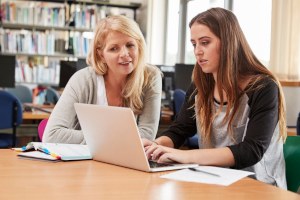 Betreuung wissenschaftlicher Arbeiten Eine ältere Person sitzt neben einer jüngeren Person, die vor sich einen geöffneten Laptop hat und etwas notiert. Die ältere Person erklärt etwas.