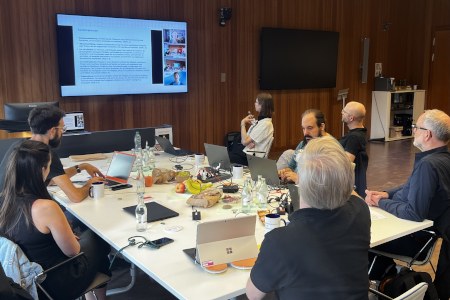 Eine Gruppe von mehreren Personen sitzen in einem Seminarraum um einen Tisch und schauen nach vorn auf eine Leinwand, auf der weitere Teilnehmende online dazugeschaltet sind.