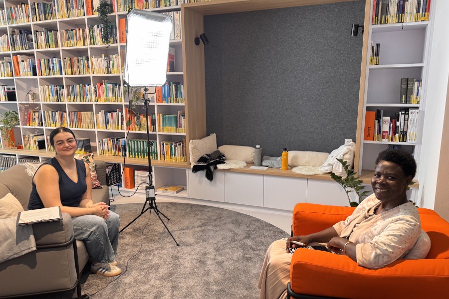 Interview mit Dr. Stella Kanyesigye Zwei Frauen sitzen sich gegenüber, im Hintergrund ist ein großes Bücherregal sowie ein Scheinwerfer zu sehen, der auf die Frau rechts gerichtet ist.