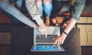 Drei Personen sitzen vor einem Laptop und zeigen mit je einer Hand auf den Bildschirm des Laptops.