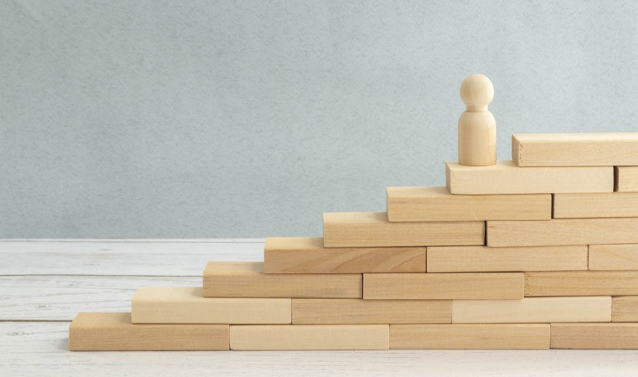 Kompetenzentwicklung Bauklötze aus Holz formen eine Treppe, auf der vorletzten Treppenstufe steht eine Spielfigur.